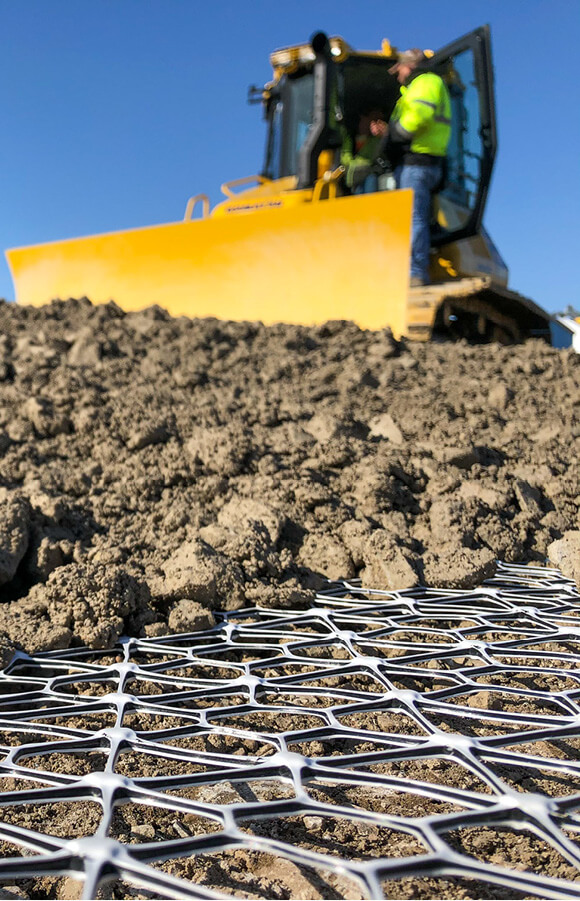 A close-up of Tensar's geogrids with soil placed on top and a construction worker and vheicle in the background